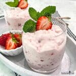 Strawberries and cream are in dessert glasses garnished with mint and a halved strawberry.