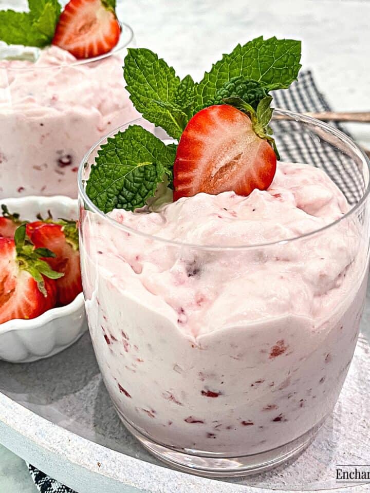 Strawberries and cream are in dessert glasses garnished with mint and a halved strawberry.