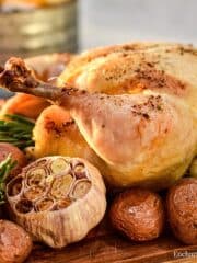 A roasted whole chicken with roasted small red potatoes and whole bulbs on a wooden cutting board garnished with fresh green rosemary.