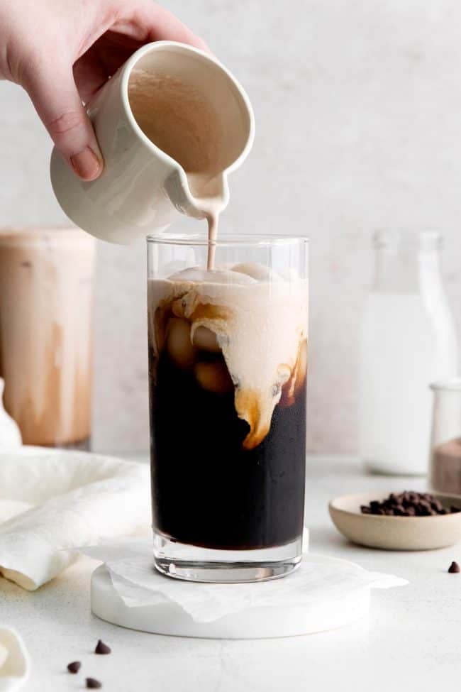 A hand pouring chocolate cream from a small pitcher into a tall glass of iced coffee.