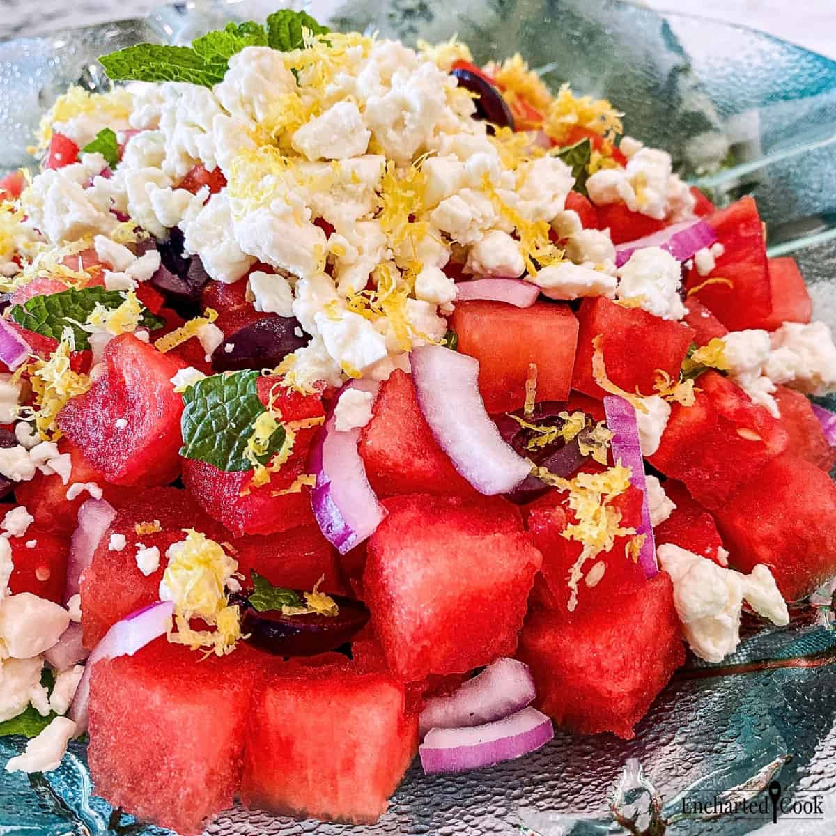 Close view of a salad of watermelon, kalamata olives red onion, and mint topped with feta cheese, and lemon zest in a bowl.