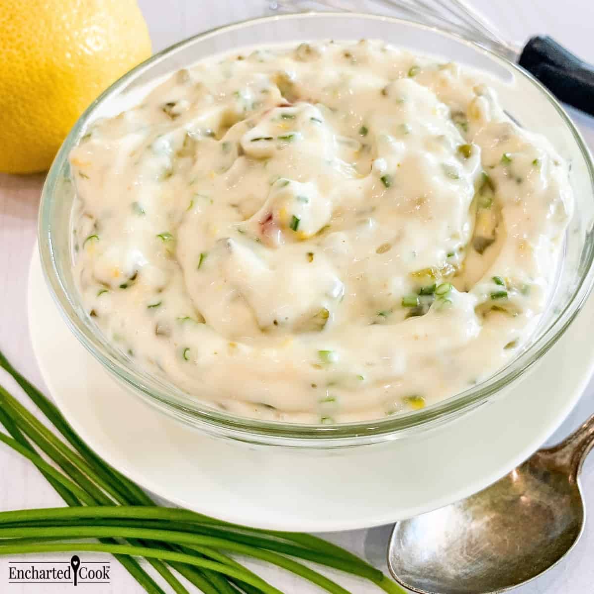 Close view of tartar sauce in a clear glass bowl on a white plate encircled by a lemon, fresh whole chives, a small whisk, and a spoon.