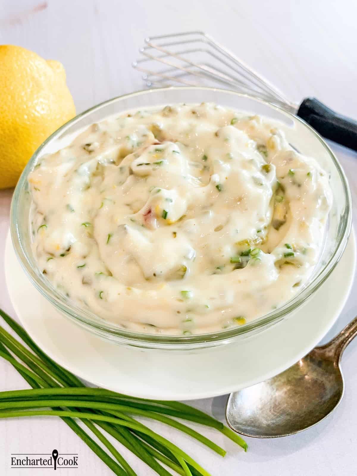 Tartar sauce in a clear glass bowl on a white plate encircled by a lemon, fresh whole chives, a small whisk, and a spoon.