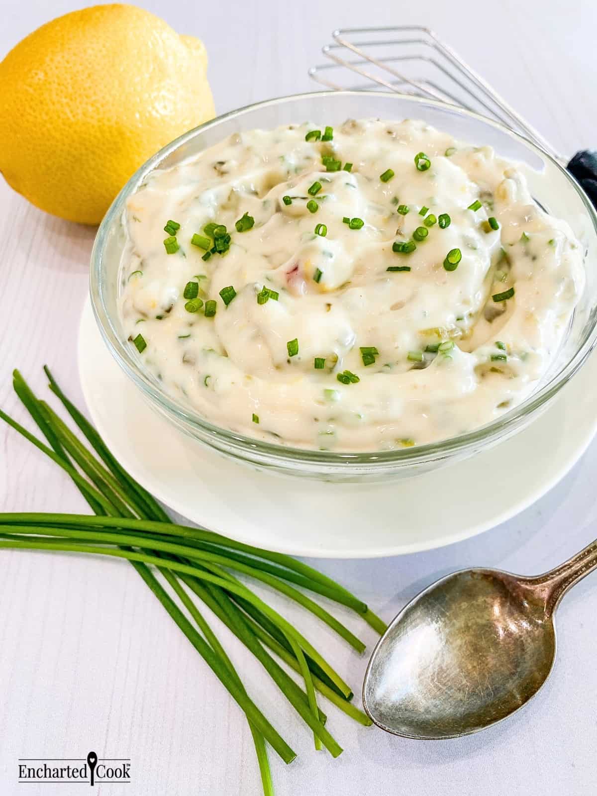 Tartar sauce in a clear glass bowl on a white plate encircled by a lemon, fresh whole chives, a small whisk, and a spoon.