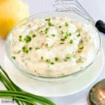 Close view of tartar sauce in a clear glass bowl on a white plate encircled by a lemon, fresh whole chives, a small whisk, and a spoon.