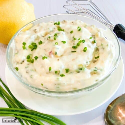 Close view of tartar sauce in a clear glass bowl on a white plate encircled by a lemon, fresh whole chives, a small whisk, and a spoon.