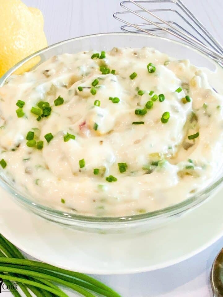Close view of tartar sauce in a clear glass bowl on a white plate encircled by a lemon, fresh whole chives, a small whisk, and a spoon.