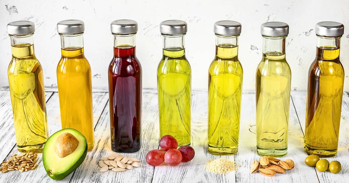 Seven bottles of different cooking oils of various clarities and colors with the fruit or seeds from which they are derived.