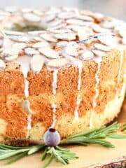 An angel food cake with a snowy white glaze and pearl frosted almonds on a wooden platter with a bark edge.