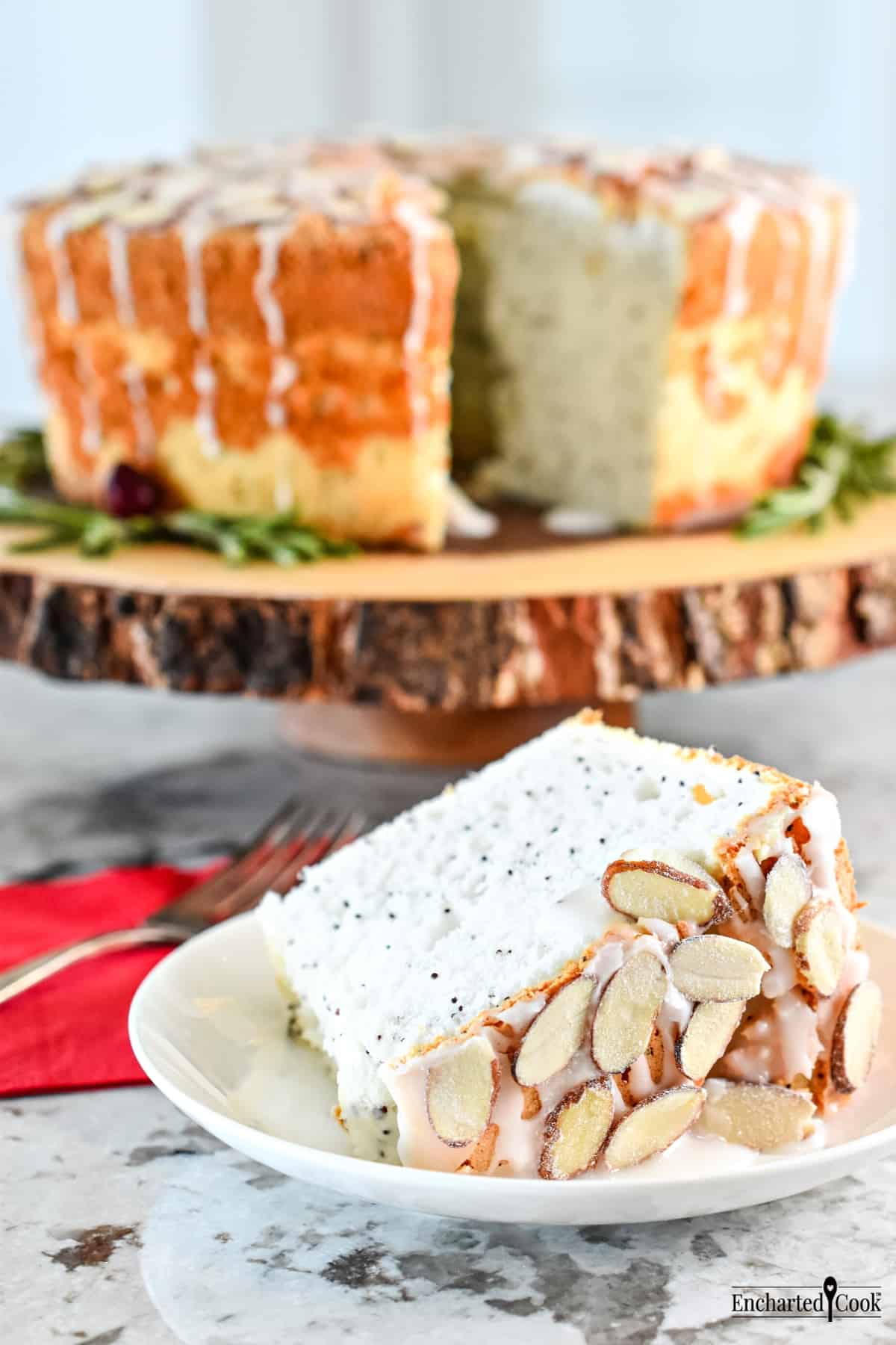 A slice of angel food cake with a snowy white glaze and pearl frosted almonds with the cut cake in the background.