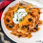A serving of a casserole of red enchilada sauce, shredded chicken, cheddar cheese, and corn tortillas on a white plate with a fork.
