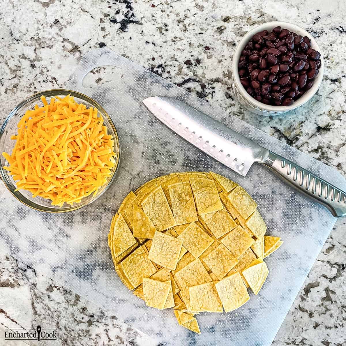 A transparent white cutting board with corn tortillas cut into bite sized pieces, drained and rinsed canned black beans, and shredded cheddar cheese.