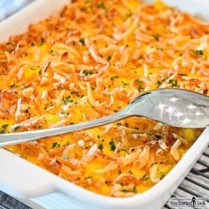 Cheesy topped casserole in a white oblong baking dish on a wire rack with a serving spoon.