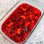 Fruited red Jello salad is in a flat clear glass baking dish.