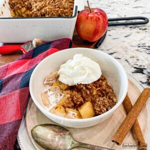 A bowl of cooked sliced apples with a crisp streusel topping and whipped cream.