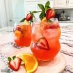 A pink and fruity sangria in a glass with ice, garnished with sliced orange and a halved strawberry.