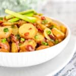 A German Potato Salad of sliced potatoes, green onions, and bacon bits dressed in a sweet and sour warm dressing in a white bowl.
