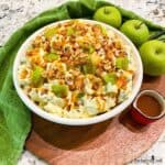 Apple and Snickers Salad in a white bowl surrounded by green apples and a small pitcher of caramel sauce.