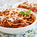 Italian Meat Sauce mixed through cooked spaghetti in a white bowl garnished with parmesan cheese and a sprig of fresh basil.