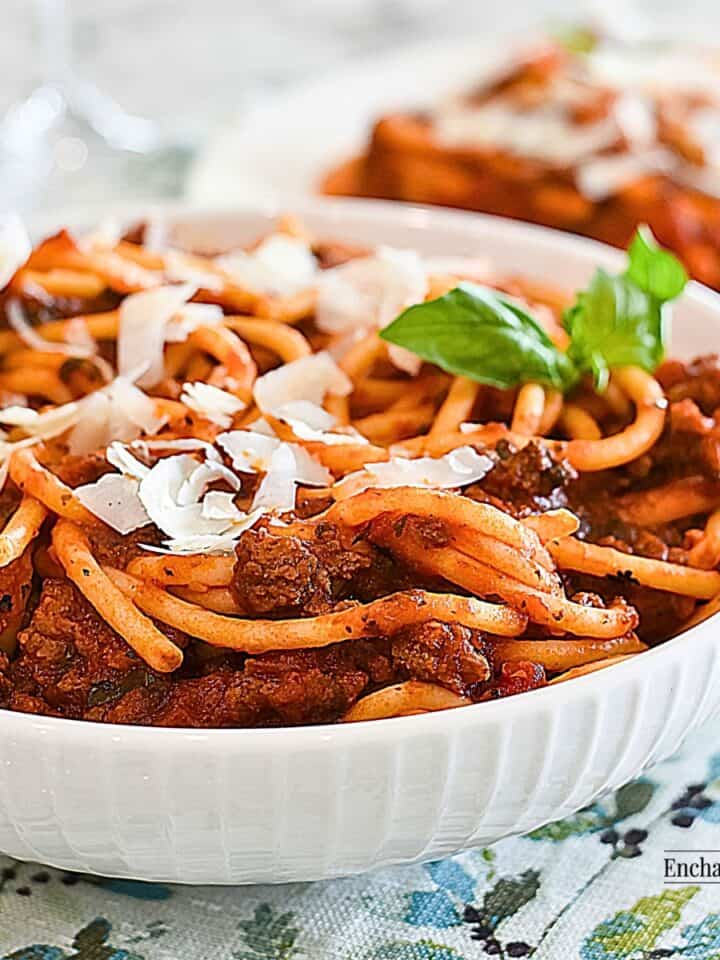 Italian Meat Sauce mixed through cooked spaghetti in a white bowl garnished with parmesan cheese and a sprig of fresh basil.