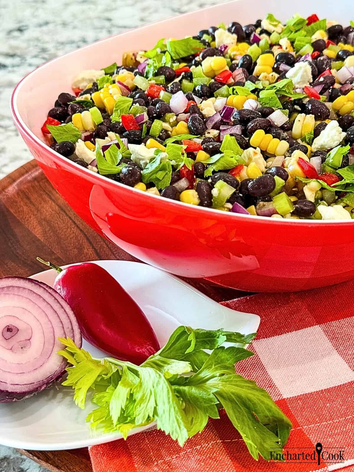 Black Bean Salad with Corn in a red and white bowl with a cut red onion, red pepper, and celery leaves on a small white plate.