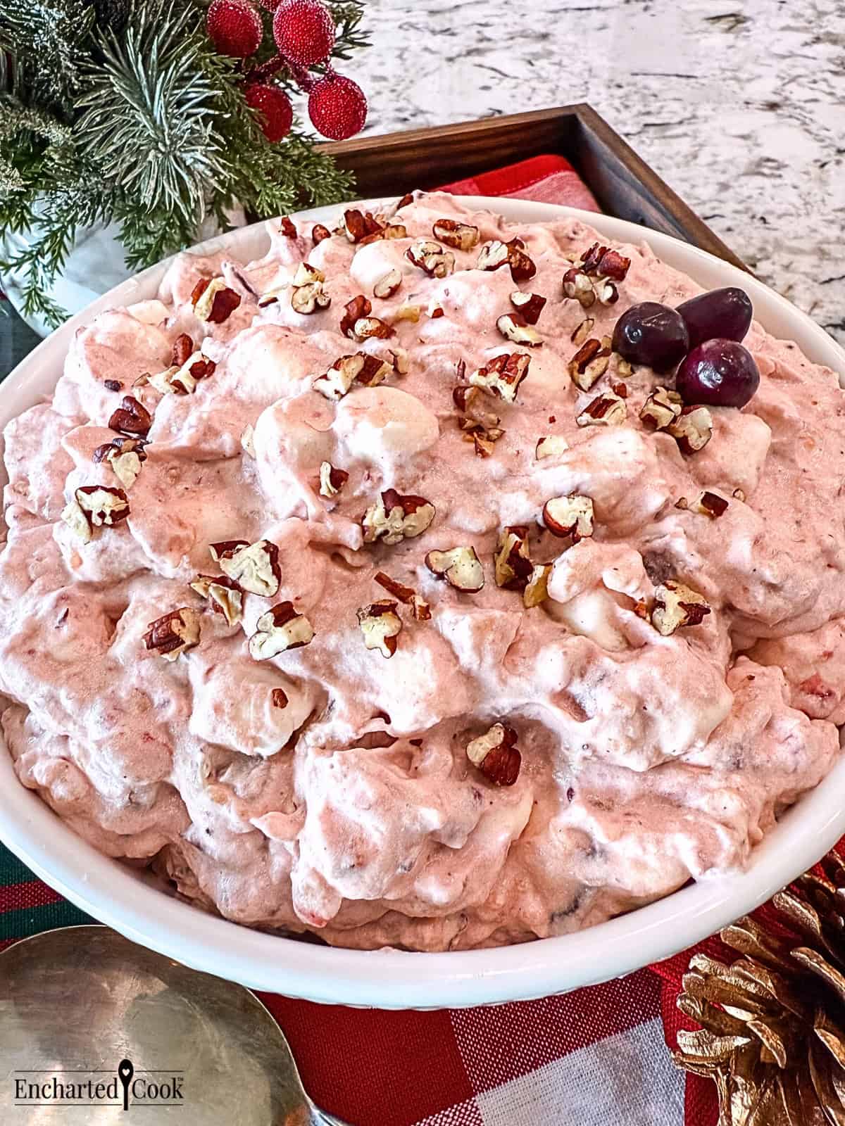 Pink Cranberry Fluff garnished with chopped pecans in a white bowl surrounded by holiday decorations.