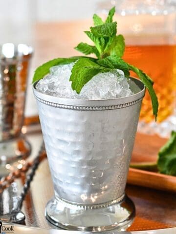 Mint leaves, a decanter of Kentucky bourbon, and a mint julip in a mint julip cup on a silver tray.