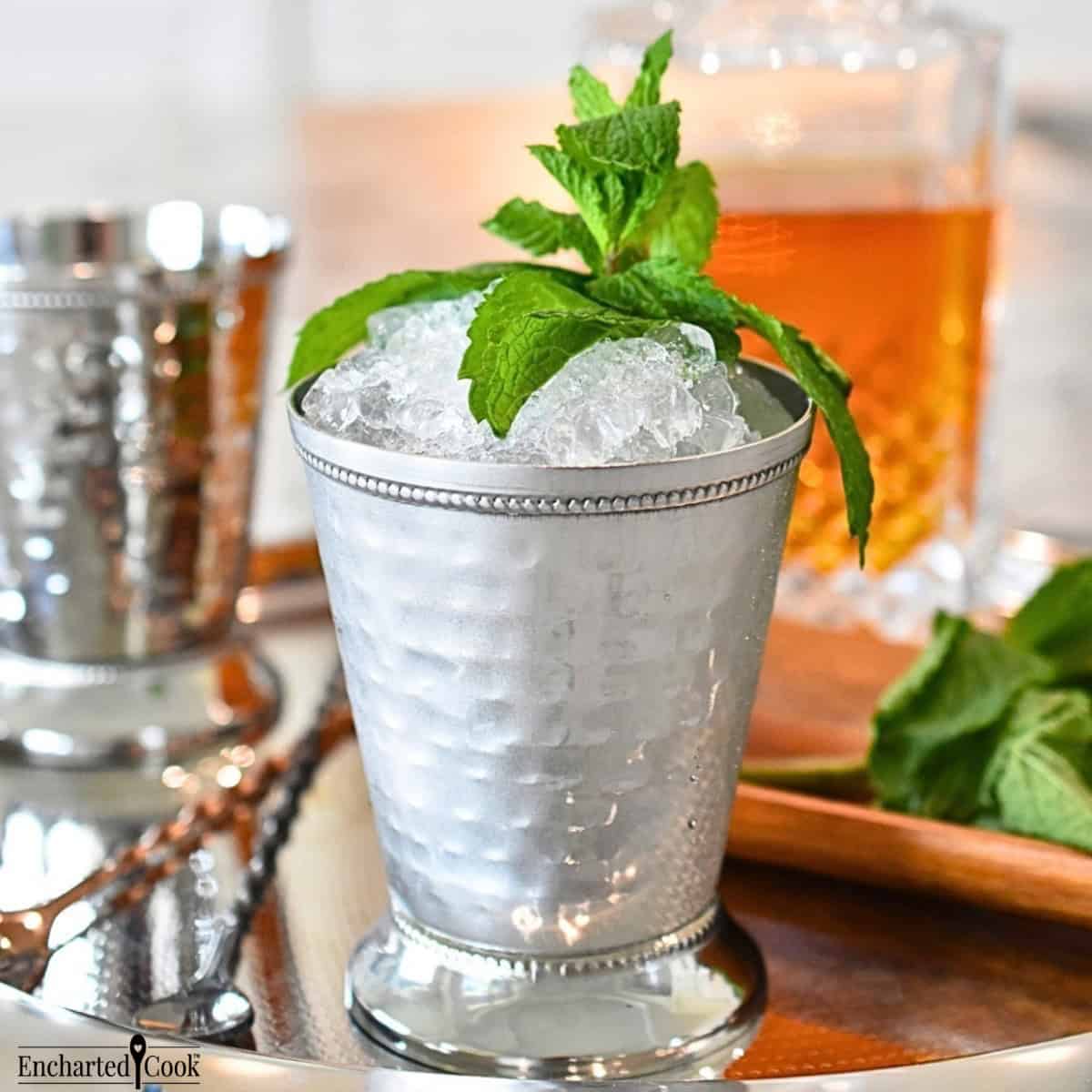 Mint leaves, a decanter of Kentucky bourbon, and a mint julip in a mint julip cup on a silver tray.