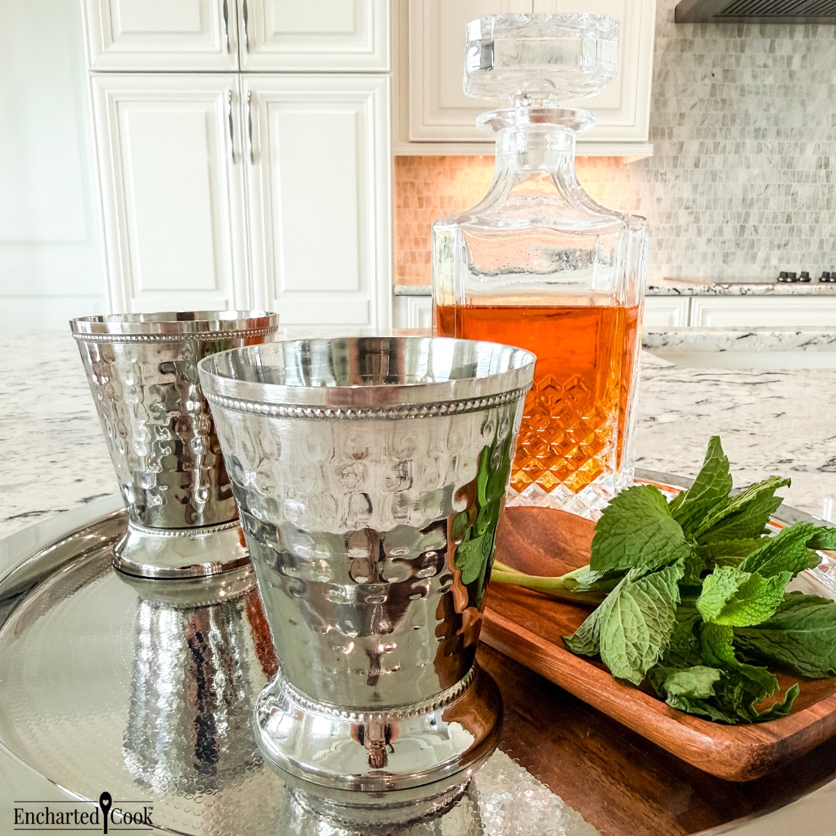 Two hammered silver colored metal julep cups on a silver tray with a decanter of bourbon and fresh mint.