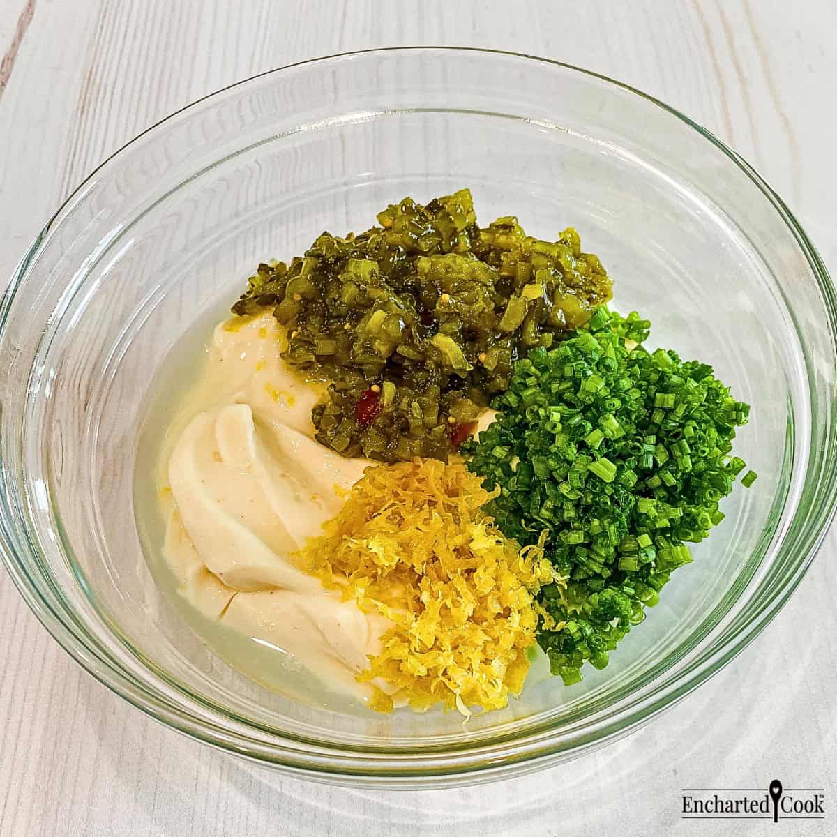 The ingredients for tartar sauce in a clear glass bowl: Miracle Whip, sweet pickle relish, fresh chopped chives, grated lemon zest, and fresh lemon juice.