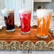 Three iced sodas, cola, fruit punch, and orange, in tall glasses are topped with Vanilla Sweet Cream Cold Foam.