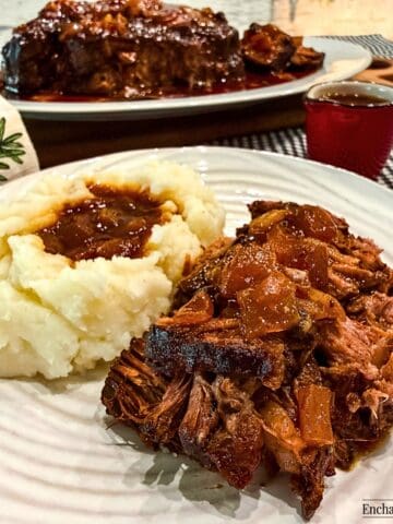 Slow cooker barbecued chuck roast is shredded and served with mashed potatoes and jus poured over the meat and the potatoes.